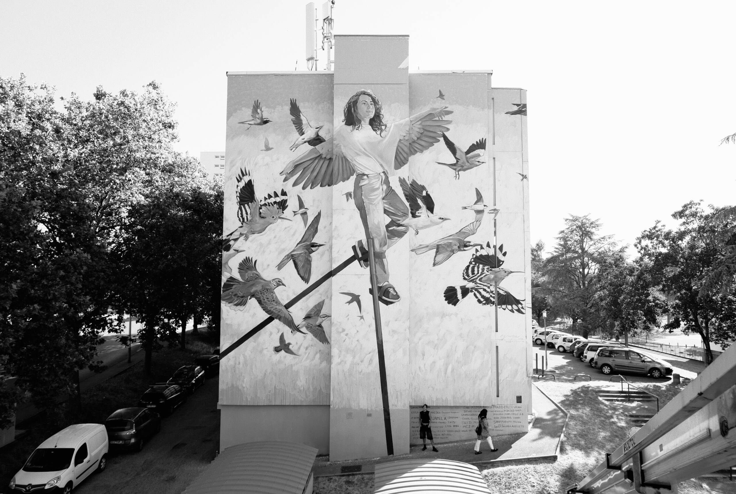 Agrume X Anamorphose Vénissieux Fresque murale en extérieur sur façade d'immeuble aux Minguettes à Vénissieux. Réalisée par Anamorphose et l'artiste Agrume. Pour Alliade Habitat. Représente une femme et des oiseaux sur fond coloré bleu et orange.