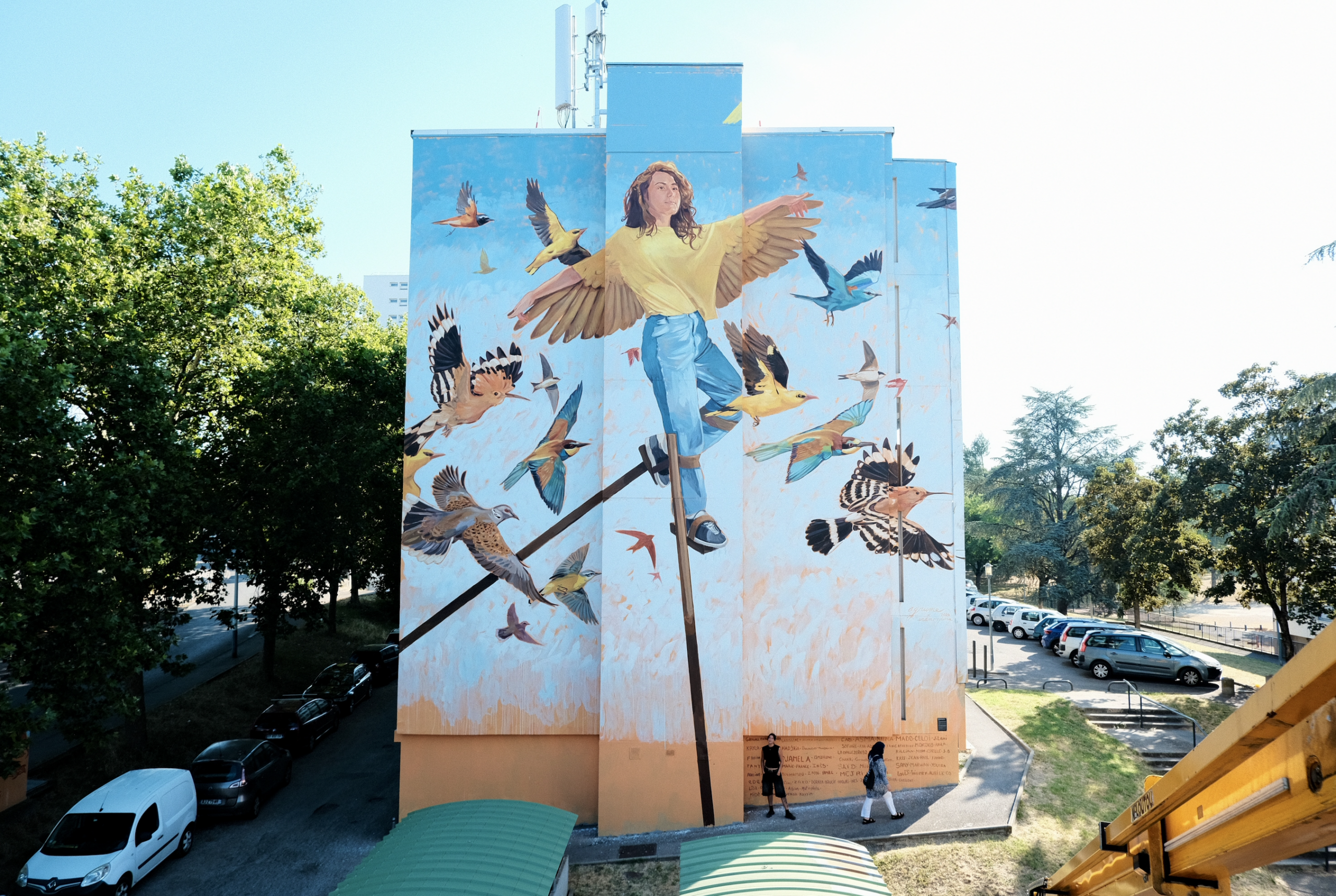 Agrume X Anamorphose Vénissieux Fresque murale en extérieur sur façade d'immeuble aux Minguettes à Vénissieux. Réalisée par Anamorphose et l'artiste Agrume. Pour Alliade Habitat. Représente une femme et des oiseaux sur fond coloré bleu et orange.